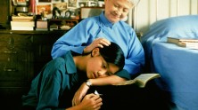 an older Asian woman with a book in her lap, stroking the hair of a kneeling younger woman with her head on the book